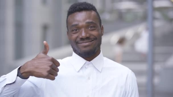 Close-up de homem afro-americano sorridente em camisa branca olhando para a câmera com o polegar para cima. Homem empresário ou gerente satisfeito com seu trabalho . — Vídeo de Stock