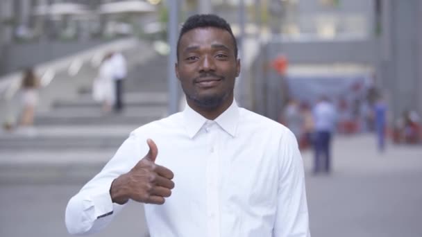 Ritratto ravvicinato di un bell'uomo afroamericano sorridente in camicia bianca che guarda la macchina fotografica con il pollice alzato mentre è in piedi sulla strada della città. Uomo d'affari o manager al lavoro — Video Stock