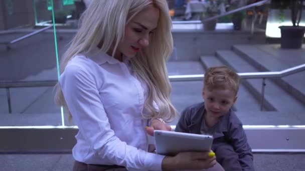 Young pretty businesswoman showing something to the small boy on her tablet. Mother entertaining her son on the backkground of the city street. — Stock videók