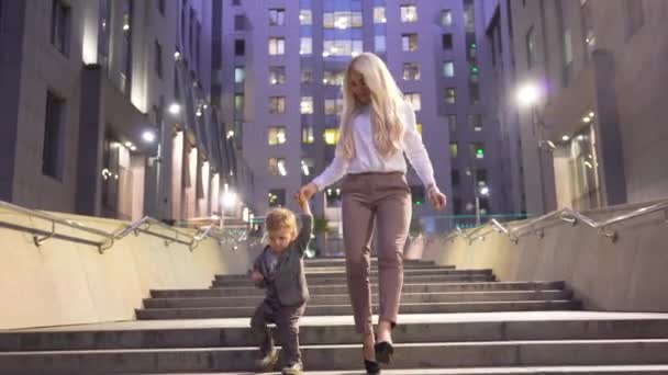 Привлекательная уверенная женщина в белой куртке и ее маленький сын, спускающийся по лестнице по городской улице. Беззаботный ребенок бежит к камере. Леди-босс проводит время со своим ребенком — стоковое видео
