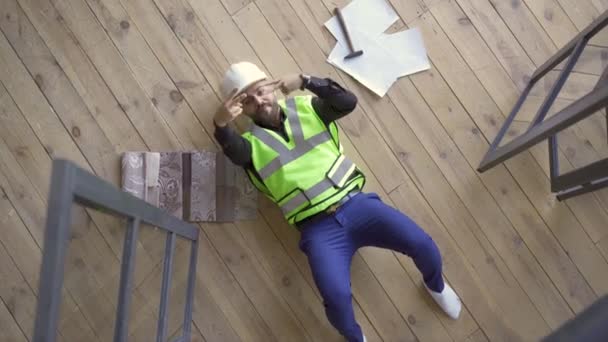 戴着头盔和绿色夹克的快乐微笑的建造者躺在地板上，展示着胜利的手势，抬头一看。职业概念。那家伙下班后休息顶部视图. — 图库视频影像