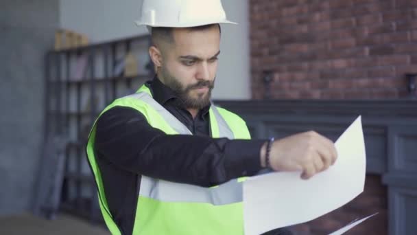 Constructor en el casco y chaqueta verde examinando dibujos. Profesional en el trabajo. Hombre mirando a través de papeles . — Vídeos de Stock