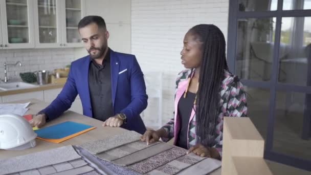 Pareja multicultural eligiendo tapicería y estilo de muebles. Hombre caucásico y mujer afroamericana decorando su casa. Diseño de interiores . — Vídeos de Stock