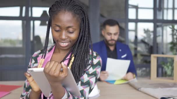 Mujer afroamericana mirando fotos en la tableta en primer plano, mientras que el diseñador masculino comprobar papeles en el fondo. Concepto de diseño de interiores — Vídeos de Stock