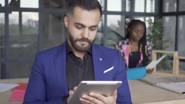 Hombre caucásico mirando fotos en la tableta en primer plano, mientras que el diseñador femenino afroamericano comprobar papeles en el fondo. Concepto de diseño de interiores — Vídeo de stock