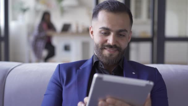 Hombre barbudo sonriente en traje azul sentado en casa trabajando con su tableta. Ocio en interiores. El hombre de negocios descansando después del trabajo. Gadget adicción . — Vídeos de Stock