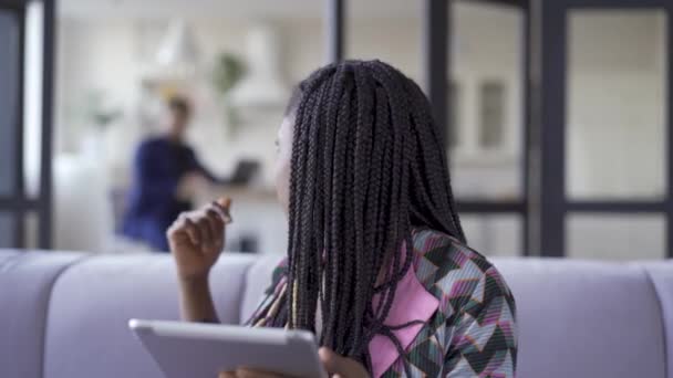 Junge hübsche afrikanisch-amerikanische Frau mit Afro-Zöpfen sitzt zu Hause und arbeitet mit ihrem Tablet. das Mädchen rief ihren Mann an, um ihm ein Bild zu zeigen. Freizeit drinnen — Stockvideo