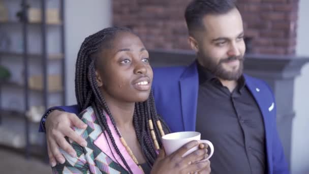 Afroamerikansk kvinna och kaukasiska man tittar på fönstret och ler. Lyckligt par med en kopp te eller kaffe tittar bort. — Stockvideo
