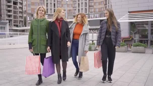 Чотири красиві подружки, що ходять по вулиці, розмовляють щасливо. Чотири модні жінки з сумками, що ходять по місту. Дозвілля разом. Дівчата насолоджуються покупками — стокове відео
