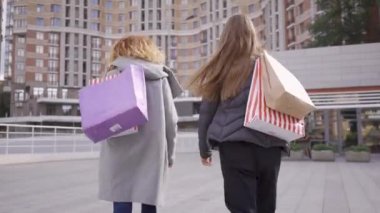 Alışveriş çantaları ile sokakta yürüyen iki güzel kız arkadaş geri görünümü konuşma ve gülümseyerek. Kaygısız yaşam tarzı. Alışverişakolik