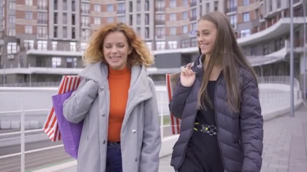 Två vackra kaukasiska flickvänner gå på gatan med shoppingväskor. Två mode kvinnor chatta och dela nyheter framåt. Bekymmerslös livsstil. Shopaholism — Stockvideo