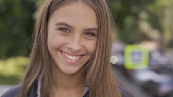 Retrato de cerca de una joven mujer caucásica con el pelo largo sonriendo felizmente mirando a la cámara en la calle de la ciudad. Emociones, felicidad, buen humor — Vídeos de Stock