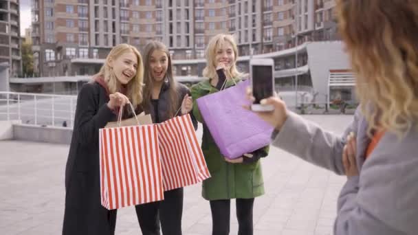 Три счастливые девушки с сумками для покупок позируют для своего четвертого друга, фотографируя. Четыре модных женщины в городе. Девушки наслаждаются покупками. Шопаголизм . — стоковое видео