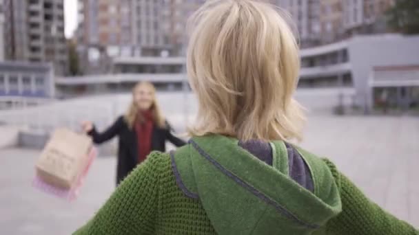 Twee blonde Kaukasische meisjes ontmoeten elkaar op straat na het winkelen. Twee mode vrouwen knuffelen, ze blij om elkaar te zien. Zorgeloze levensstijl. Shopaholism. — Stockvideo