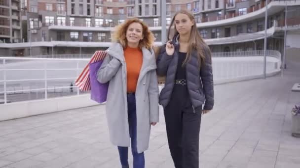 Twee gelukkige Kaukasische meisjes wandelen met boodschappentassen. Twee mode vrouwen chatten en delen van nieuws vooruit. Zorgeloze levensstijl. Shopaholism. — Stockvideo