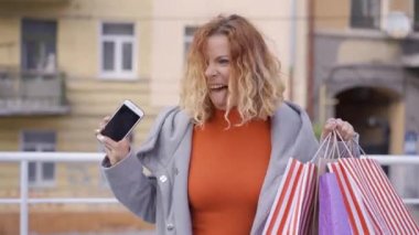 Heyecanlı kızıl saçlı kız elinde alışveriş çantaları ve cep telefonu ile havada yürüyor. Yetişkin kadın onun alımları memnun. Moda konsepti.