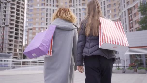 Vue arrière de deux copines marchant dans la rue avec des sacs à provisions. Style de vie insouciant. Shopaholisme . — Video