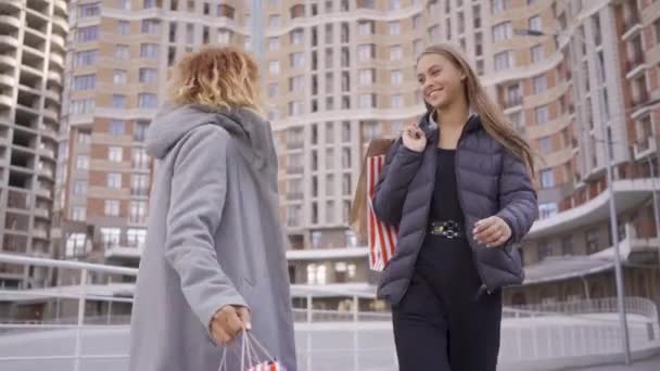 Twee gelukkige mooie meiden lopen op straat met boodschappentassen, praten en glimlachen. Zorgeloze levensstijl. Shopaholism. — Stockvideo