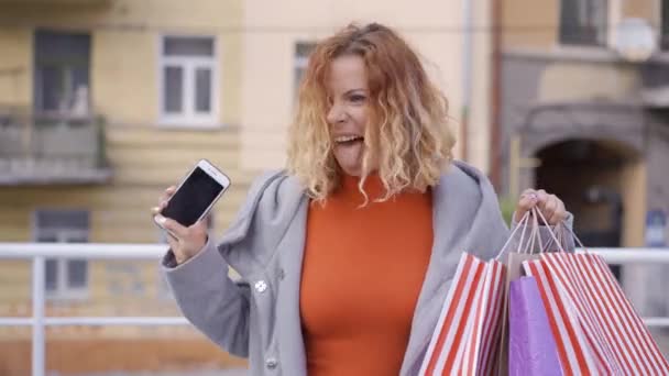 Fille aux cheveux roux excitée avec des sacs à provisions et un téléphone portable dans les mains marchant sur les airs. Femme adulte satisfaite de ses achats. Concept de mode . — Video