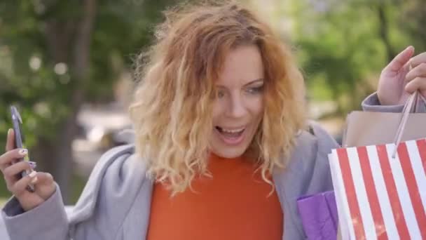 Close-up of red-haired shopaholic with shopping bags and cellphone in hands jumping and rising hands up in victory gestures on the street. Leisure of city dweller. Fashion concept. — Stock Video