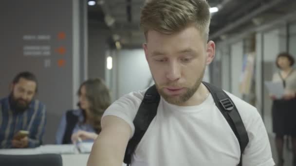 Beau jeune homme en t-shirt blanc travaillant avec un ordinateur portable assis à la table dans le bureau moderne. gestion du travail terminé. Lieu de travail . — Video