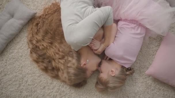 집에서 푹신한 카펫에 누워 빨간 드레스에 행복 한 백인 어머니와 그녀의 작은 딸의 상단보기. 여자와 여자는 집에서 쉬고 미소. 행복한 가족. — 비디오