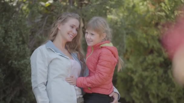 Joven madre rubia sosteniendo a su linda hijita en las manos en el parque. Mujer caucásica pasando la noche de otoño con el niño vestido con chaqueta roja . — Vídeo de stock