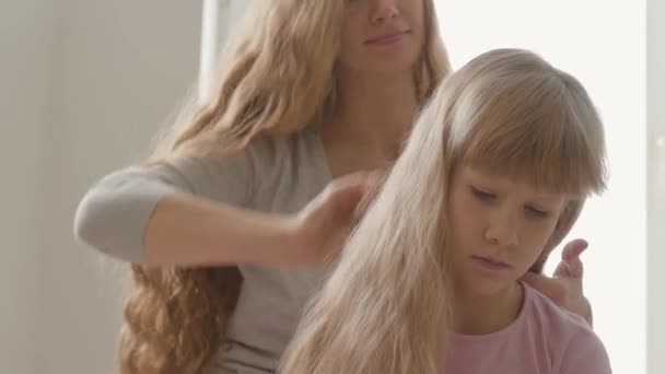 Κοντά χέρια της νεαρής γυναίκας που χτενίζει τα μαλλιά της ξανθιάς κόρης της. Η ιδέα της μητρότητας, της παιδικής ηλικίας, ενός γονέα. Αναψυχή σε εσωτερικούς χώρους. Ευτυχισμένη οικογένεια — Αρχείο Βίντεο