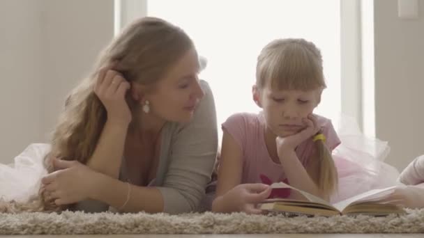 Unbeschwerte blonde kaukasische Mutter und kleine Tochter auf dem Bett liegend. hübsche Frau, die dem kleinen Mädchen den Inhalt des Buches erklärt. Kindererziehung. — Stockvideo