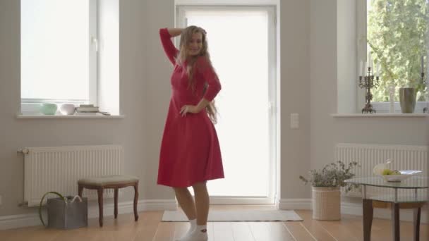 Krásná usmívající se bělošská žena s dlouhými kudrnatými vlasy v červených šatech tančících doma. Atraktivní mladá dívka, která se baví uvnitř. — Stock video