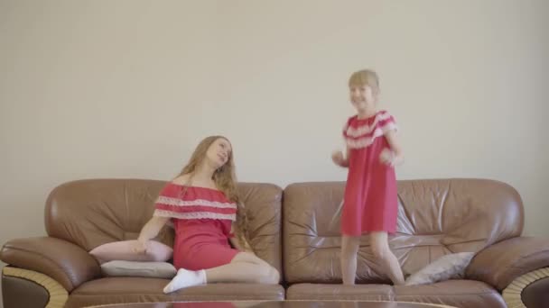 Tired young woman looking at the little cute girl jumping on the coach. Mother and daughter in the same red dresses playing in the evening at home. — Stock Video