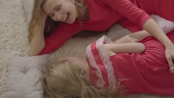 Top uitzicht op mooie jonge vrouw en blond meisje op het zachte tapijt en zachte kussens. Kaukasische moeder en dochter in rode jurken uitgaven avond thuis rusten. — Stockvideo