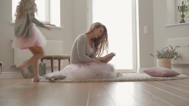 Jolie femme caucasienne en robe rouge et chemisier gris assis sur le tapis et la lecture. Petite fille joyeuse courant vers sa mère par le dos et fermant les yeux des mamans avec les mains . — Video