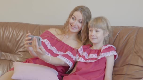 Mãe branca loira feliz e filha pequena nos mesmos vestidos sentados no sofá tomando selfie usando celular em casa. Conceito de maternidade, infância, um dos pais. Família feliz — Vídeo de Stock