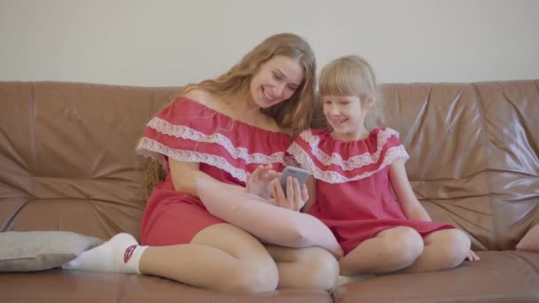 Feliz madre rubia caucásica y su hija pequeña en los mismos vestidos sentados en el sofá viendo fotos en el teléfono celular en casa. Concepto de maternidad, infancia, un padre. Familia feliz — Vídeos de Stock