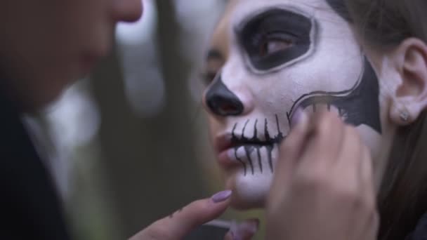 Frauenhände tragen mit dem kleinen Pinsel in Großaufnahme schwarze Farbe auf das Gesicht des Mädchens auf. Junge Leute bereiten sich auf die Feier von Halloween vor und malen sich den Totenkopf ins Gesicht. Professionelles Make-up — Stockvideo
