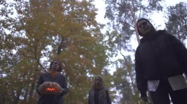 Cadılar Bayramı kostümlü erkek ve iki genç kadın sonbahar parkında ciddi korkunç yüzlerle kurbana bakıyorlar. Büyük bir bıçak gösteren, öldürecek olan kız, balkabağını tutan adam. Alt görünüm