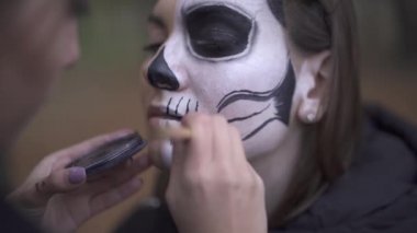 Arkadaşlarının yüzüne Cadılar Bayramı makyajı yapan beyaz bir kızın yakın çekimi. İki gotik kadın azizler gecesi kutlamalarıiçin hazırlanıyor. Üzerinde korkunç bir vücut-sanat resmi ile Kadın yüzü.