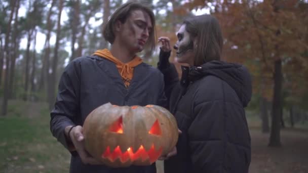 Uomo e donna in costumi di Halloween con make-up guardando nella fotocamera con gravi volti spaventosi nel parco autunnale. L'uomo che tiene una grande zucca con dentro una candela accesa — Video Stock