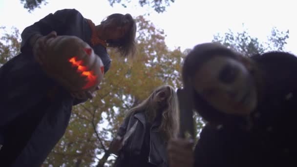 Homme et deux femmes en costumes d'Halloween regardant la victime avec de sérieux visages effrayants dans le parc d'automne. La fille qui montre un gros couteau, va tuer, le gars qui tient de la citrouille. Vue du bas — Video