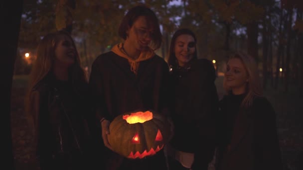 Knappe man houdt Halloween pompoen met een licht binnen, drie meisjes staan in de buurt. Mensen met enge make-up op gezichten die glimlachen en dan naar de camera gaan met kwade looks — Stockvideo