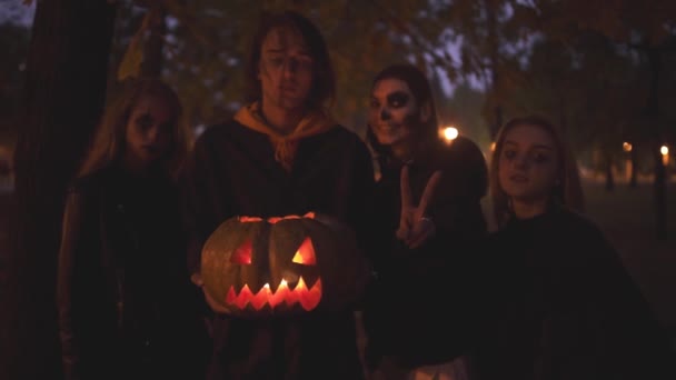 Τέσσερις λευκοί φίλοι γιορτάζουν τη νύχτα των Αγίων πάντων. Ομάδα νέων ανθρώπων με ένα τρομακτικό αποκριάτικο σπίτι στο νυχτερινό πάρκο κρατώντας μια κολοκύθα με ένα κερί μέσα. — Αρχείο Βίντεο