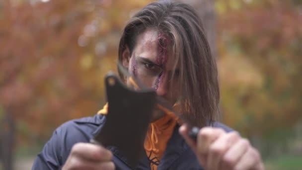 Un hombre guapo con una gran cicatriz fea en la cara demostrando dos cuchillas, frotándose entre sí. El tipo con maquillaje de Halloween en la cara frotando sus cuchillos con una mirada malvada. Cambio de enfoque — Vídeos de Stock