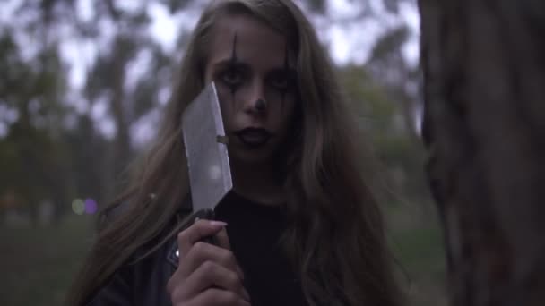 Jeune femme folle avec du maquillage de clown d'Halloween sur le visage tenant un gros couteau à la main, riant. La fille regarde avec colère dans la caméra et frappe le tronc de l'arbre avec un coupe-branches — Video