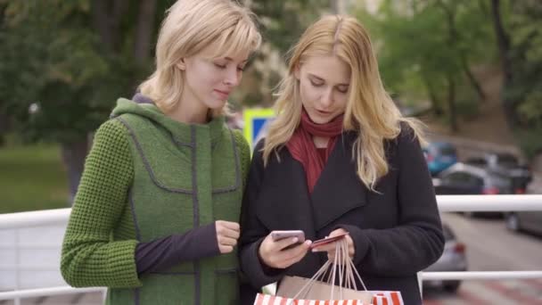 Dos hermosas novias rubias de pie en la calle con sus compras en bolsas de compras. La mujer que hace el pago en línea usando su teléfono celular y tarjeta de crédito. Shopaholism, pago fácil — Vídeo de stock