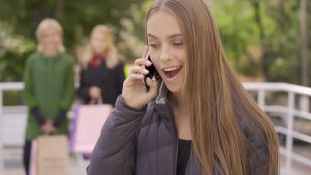 Portrait de femme caucasienne positive parlant par téléphone portable tandis que deux de ses copines l'attendent en arrière-plan. Loisirs ensemble. Les filles passent du temps dans la ville — Video