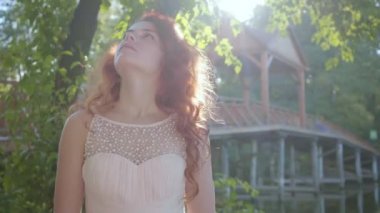 Parkta duran ve bir şey üzerinde düşünen bej elbiseli güzel kızıl saçlı kadın. Yaz güneş ışınlarında düşünceli beyaz bir kızın portresi.