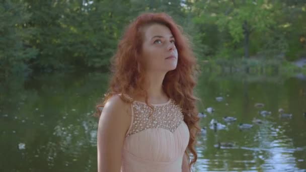 Pretty caucasian woman with red hair in beige dress standing on the bank of the lake and looking up and around. Redhead thoughtful girl resting outdoors. — Stock Video