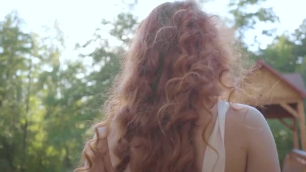 Rückansicht der schönen, schlanken Frau mit roten Haaren, die im Garten spaziert, die Kamera folgt ihr. Freizeit im Freien. Verbindung zur Natur — Stockvideo