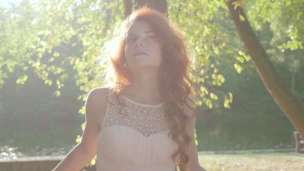 美丽的苗条的白种女人的肖像，红头发的米色礼服站在公园或花园的阳光下，望向远方。与自然的联系。特写视图 — 图库视频影像